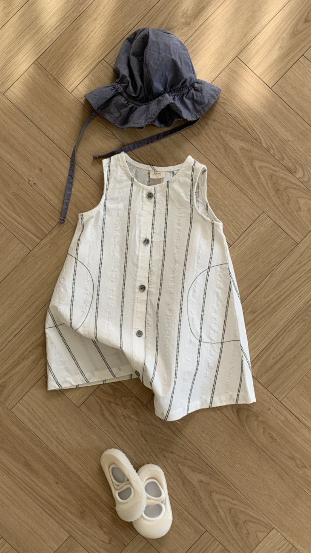 Ein auf einem Holzboden ausgebreitetes Kinderoutfit, bestehend aus einem gestreiften ärmellosen Oberteil mit Knopfleiste, einem Schlapphut aus Blau und einem einzelnen weißen Schuh von Bebe Holics Daisy ST Body Suit.