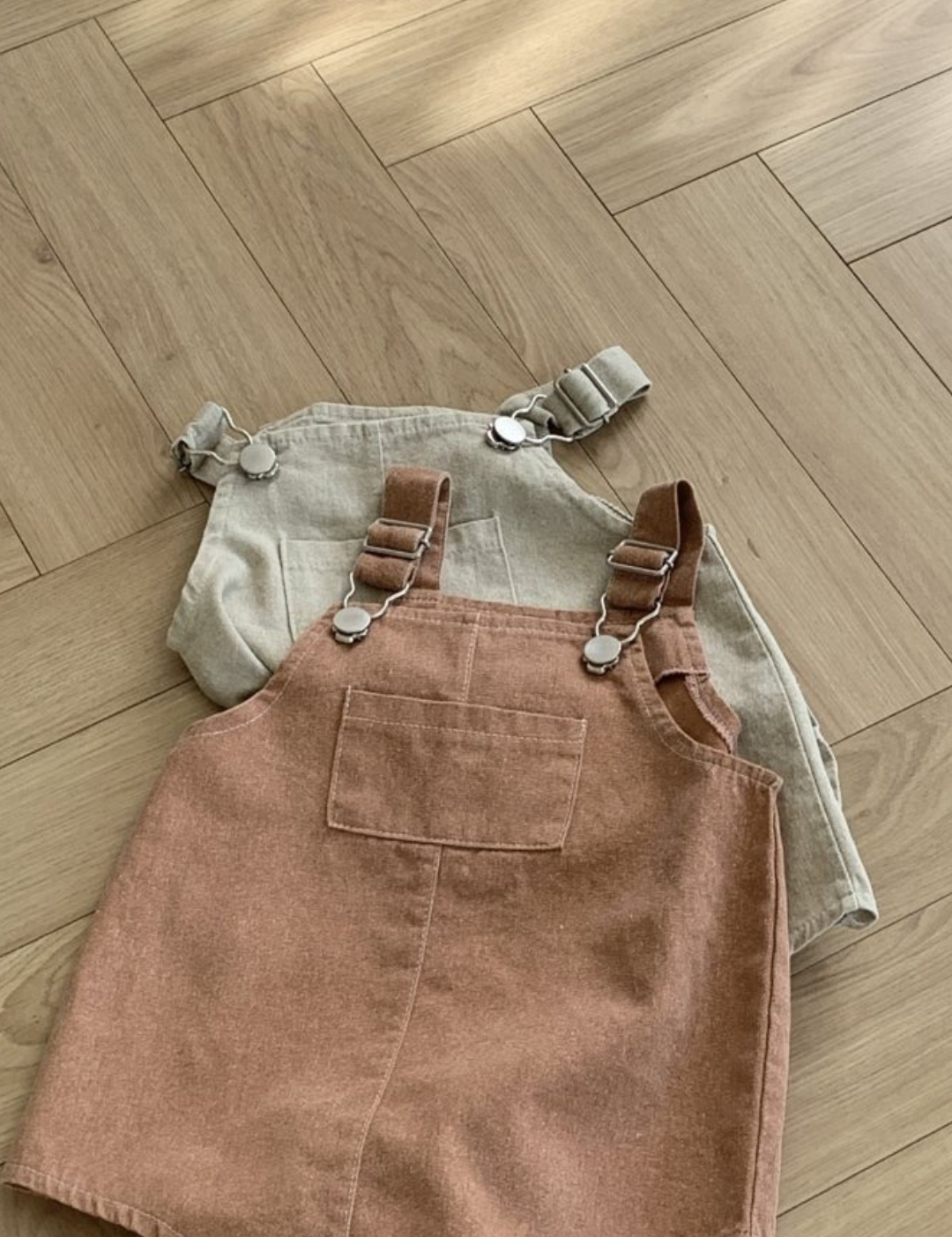 Zwei Paar Kinderoveralls liegen auf einem Holzboden. Das Paar im Vordergrund, gefertigt von Bebe Holic, ist ein Pigment Dungarees Body Suit in einem sanften Braunton mit verstellbaren Trägern und einer Vordertasche. Das Paar im Hintergrund ist hellbeige mit ebenso verstellbaren Trägern. Sonnenlicht fällt von der oberen linken Ecke ein.