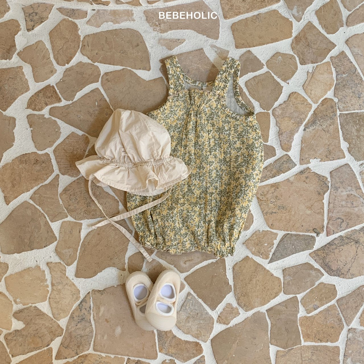 Auf einem Steinboden liegt ein Kleinkinderoutfit. Es besteht aus einem Karen-Bodysuit aus atmungsaktiver Baumwolle, einem beigen Sonnenhut mit Kinnriemen und einem Paar elfenbeinfarbener Babyschuhe. Der Markenname „Bebe Holic“ ist oben im Bild zu sehen.