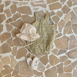 Auf einem Steinboden liegt ein Kleinkinderoutfit. Es besteht aus einem Karen-Bodysuit aus atmungsaktiver Baumwolle, einem beigen Sonnenhut mit Kinnriemen und einem Paar elfenbeinfarbener Babyschuhe. Der Markenname „Bebe Holic“ ist oben im Bild zu sehen.