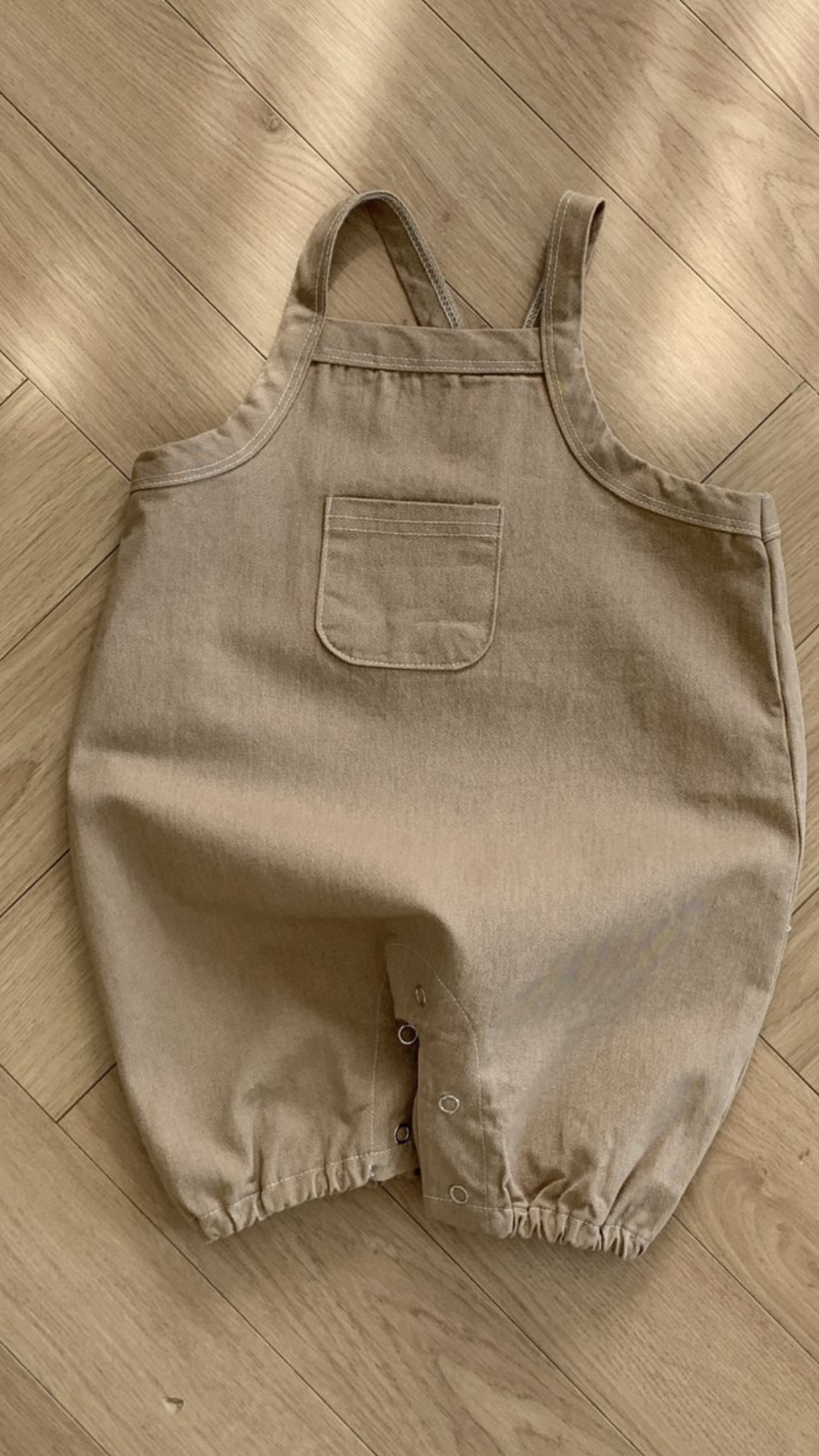 Ein Bild eines beigen Baby-Stramplers aus Twill mit Taschen von Bebe Holic, der flach auf einem Holzboden liegt. Der Strampler hat eine Vordertasche, Knopfverschlüsse im Schritt und Rüschen am Bein.