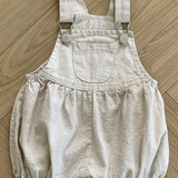 Ein beiger Baby-Strampler aus atmungsaktiver Baumwolle mit verstellbaren Trägern und Metallknopfverschlüssen auf Holzboden. Diese Bebe Holic Leinen-Latzhose mit Schnalle hat eine Vordertasche, eine geraffte Taille und locker sitzende Shorts für mehr Komfort.