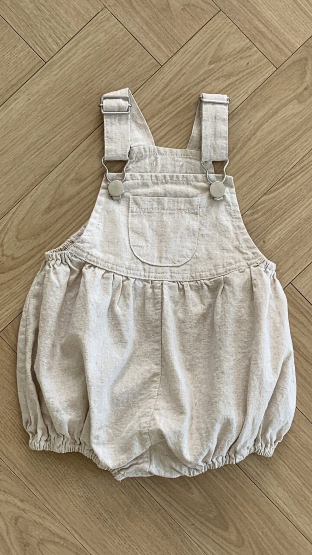 Ein beiger Baby-Strampler aus atmungsaktiver Baumwolle mit verstellbaren Trägern und Metallknopfverschlüssen auf Holzboden. Diese Bebe Holic Leinen-Latzhose mit Schnalle hat eine Vordertasche, eine geraffte Taille und locker sitzende Shorts für mehr Komfort.