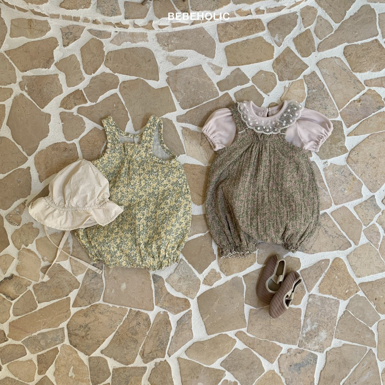 Zwei Kinderoutfits liegen auf einem Boden mit Steinmuster. Das Outfit links besteht aus einem geblümten Strampler und einem beigen Anglerhut. Das Outfit rechts besteht aus einer rosa Bluse mit Karen-Bodysuit und braunen Schuhen. Oben ist das Wort „Bebe Holic“ zu sehen.