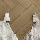 Zwei Leinen-Latzhosen mit Schnallen von Bebe Holic liegen auf einem Holzboden. Der weiße Overall ist links und hat verstellbare Träger mit Metallschnallen und geraffte Seiten. Der hellbeige Overall rechts hat ebenfalls verstellbare Träger mit Metallschnallen und geraffte Seiten und ist aus atmungsaktiver Baumwolle.