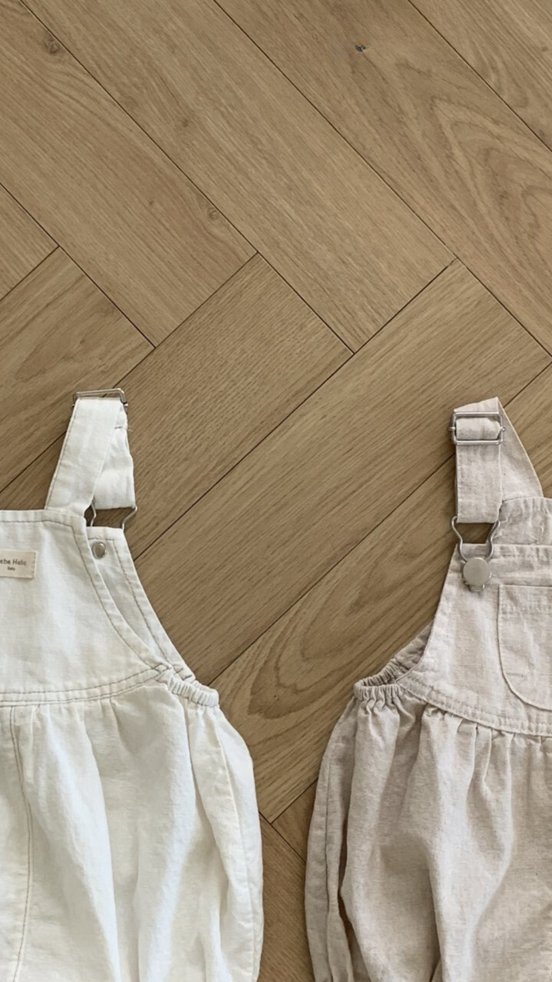 Zwei Leinen-Latzhosen mit Schnallen von Bebe Holic liegen auf einem Holzboden. Der weiße Overall ist links und hat verstellbare Träger mit Metallschnallen und geraffte Seiten. Der hellbeige Overall rechts hat ebenfalls verstellbare Träger mit Metallschnallen und geraffte Seiten und ist aus atmungsaktiver Baumwolle.