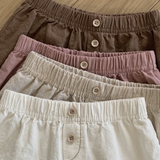 Vier Paar Bebe Holic Natural Pants mit elastischer Taille liegen auf einer Holzfläche. Die Shorts sind in verschiedenen Farben erhältlich: Braun, Mauve, Beige und Creme, von oben bis unten. Jedes Paar hat vorne in der Mitte einen Knopfverschluss – ein vielseitiges Must-have für jede Garderobe.