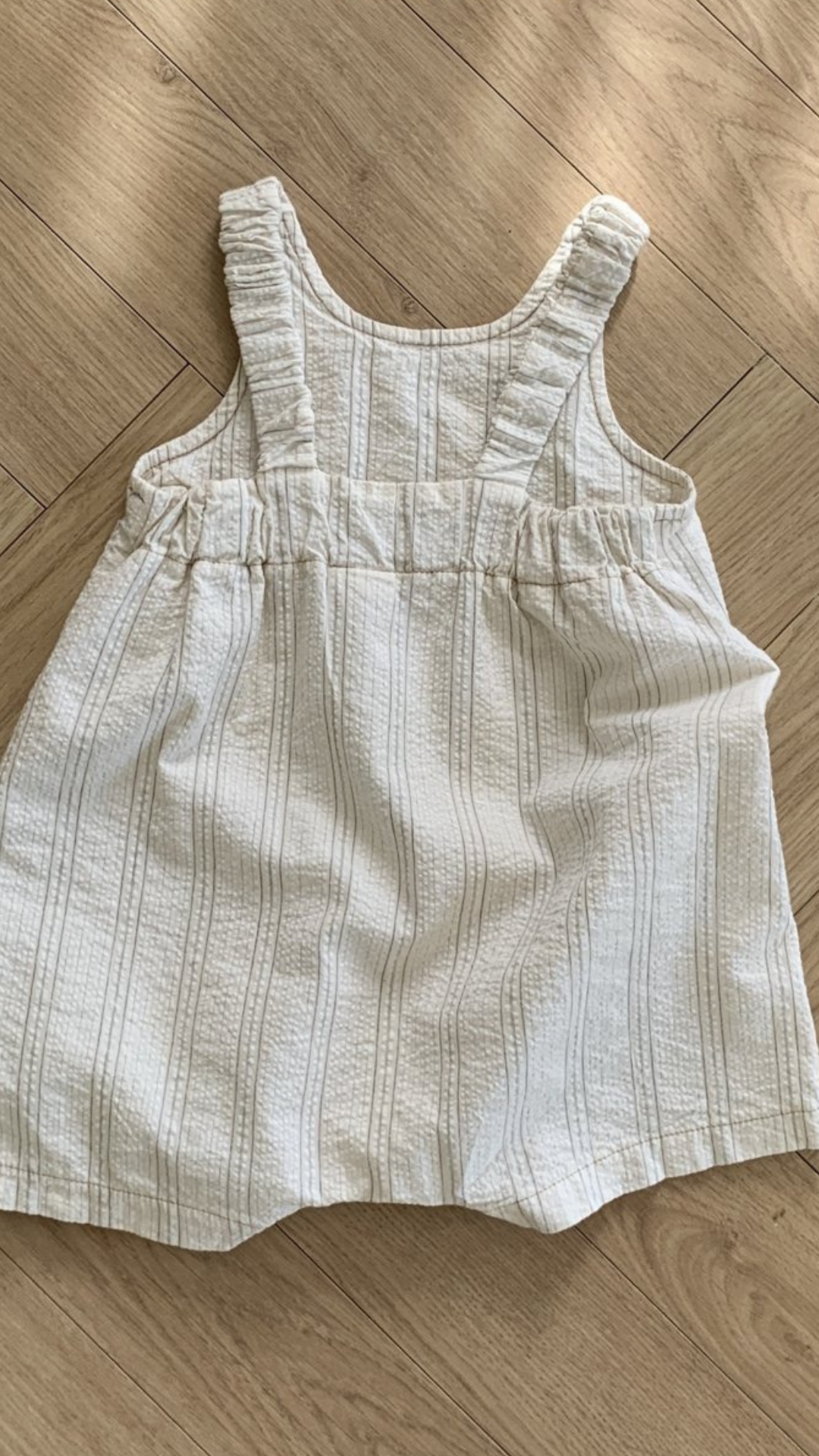 Ein ärmelloses Kleinkinderkleid von Bebe Holic Naturtöne mit dünnen Trägern und einer gerüschten Taille, das flach auf einem Holzboden liegt. Das Kleid hat ein dezentes Karomuster.