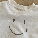 Eine Nahaufnahme eines weißen Bebe Holic Smile Tee mit einem einfachen schwarzen Smiley-Design auf der Vorderseite. Das Shirt aus weicher Baumwolle liegt flach auf einem Holzboden und das Etikett mit „Bebe“ ist am Ausschnitt teilweise sichtbar.