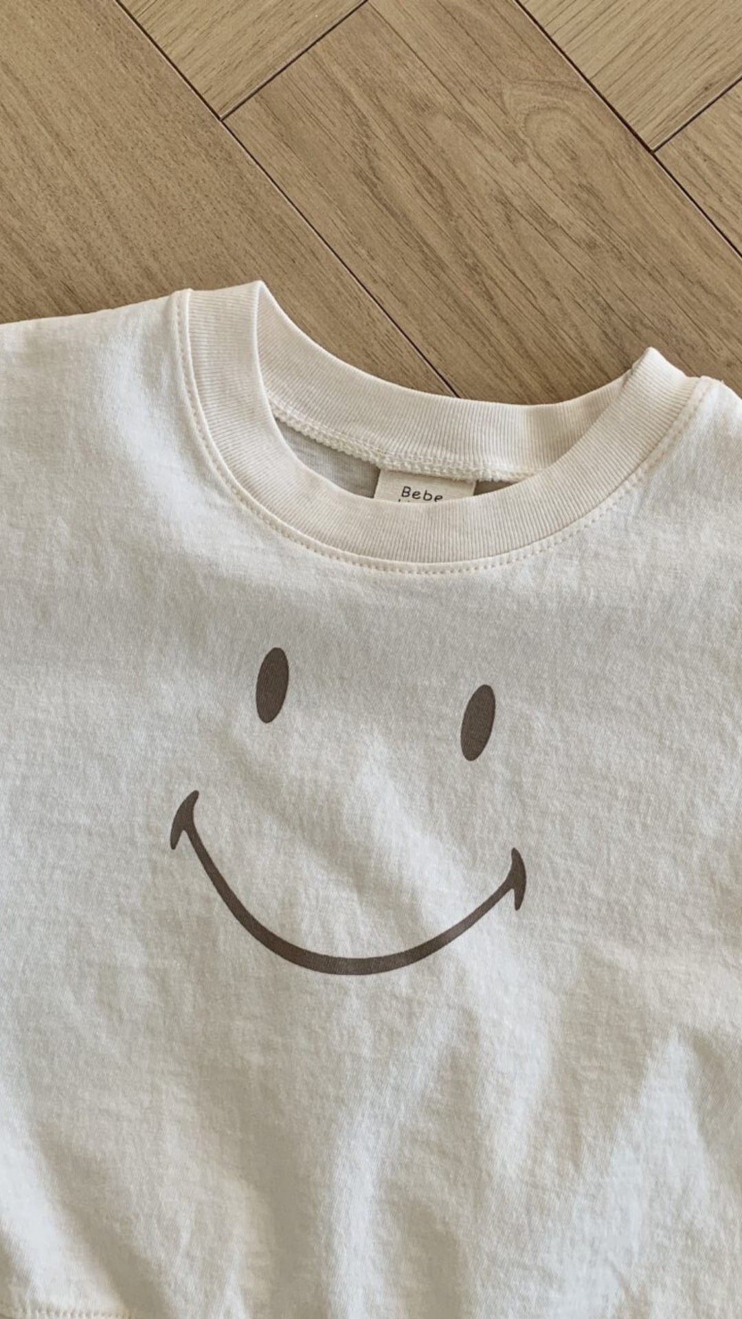 Eine Nahaufnahme eines weißen Bebe Holic Smile Tee mit einem einfachen schwarzen Smiley-Design auf der Vorderseite. Das Shirt aus weicher Baumwolle liegt flach auf einem Holzboden und das Etikett mit „Bebe“ ist am Ausschnitt teilweise sichtbar.