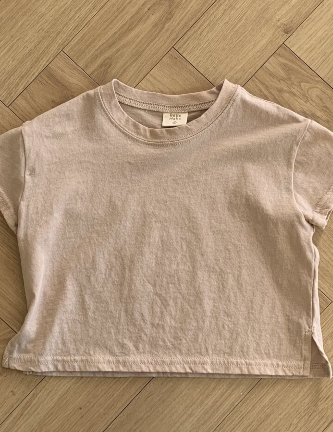 Ein Bebe Holic Basic Tee liegt flach auf einem Holzboden. Das Shirt aus hochwertiger Baumwolle hat einen runden Halsausschnitt und kleine Seitenschlitze am Saum. In der Nähe des Innenkragens ist ein Etikett zu sehen.