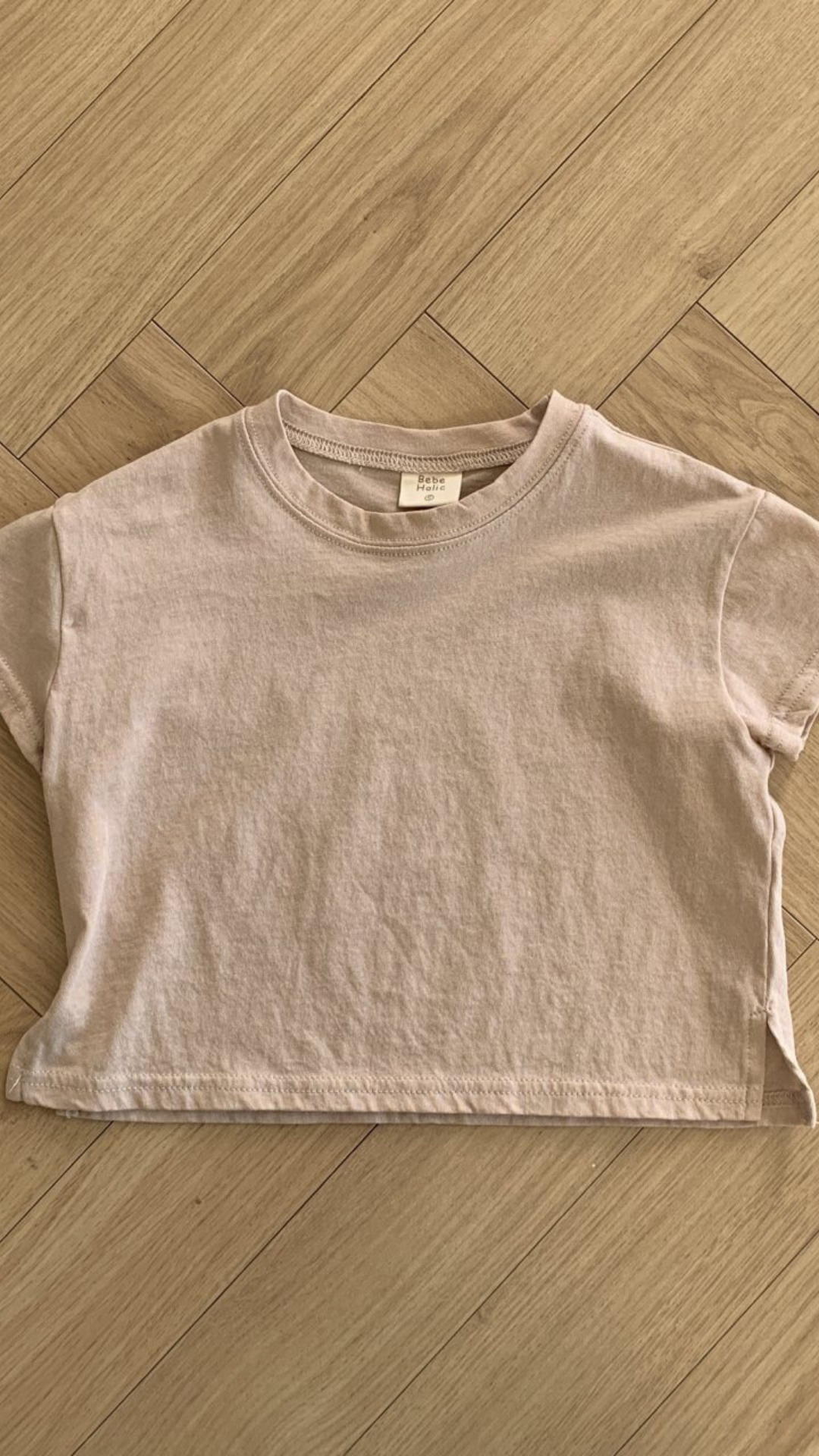 Ein Bebe Holic Basic Tee liegt flach auf einem Holzboden. Das Shirt aus hochwertiger Baumwolle hat einen runden Halsausschnitt und kleine Seitenschlitze am Saum. In der Nähe des Innenkragens ist ein Etikett zu sehen.