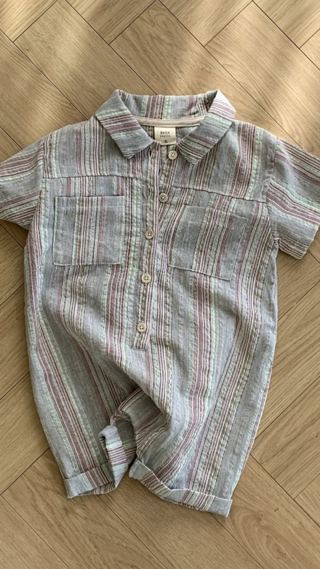 Ein Aurora-Bodysuit für Kleinkinder von Bebe Holic mit kurzen Ärmeln, Kragen und Knopfverschluss vorne liegt flach auf einem Holzboden. Der Strampler hat vertikale Streifen in Grau-, Rosa- und Weißtönen.