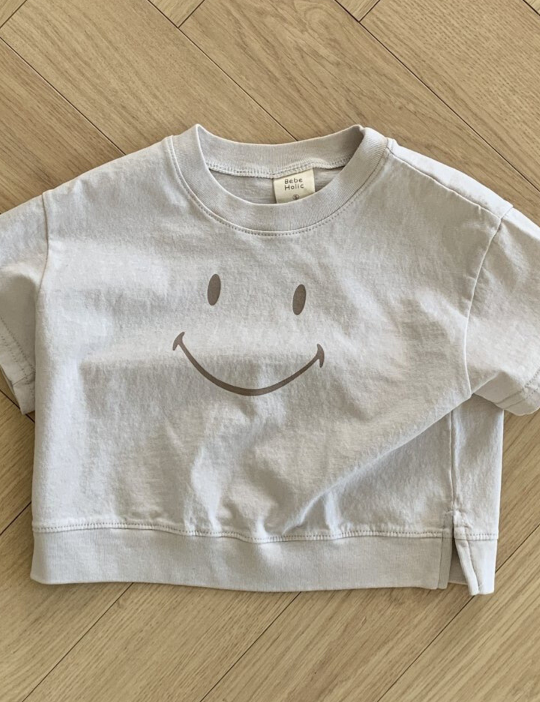 Ein weißes, kurzes T-Shirt namens Smile Tee aus der Bebe Holic-Kollektion mit einem großen Smiley auf der Vorderseite liegt auf einem hellen Holzboden mit Fischgrätenmuster. Das Smile Tee besteht aus weicher Baumwolle, hat kurze Ärmel und ein lässiges Design.