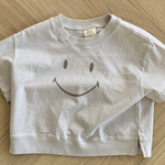 Ein weißes, kurzes T-Shirt namens Smile Tee aus der Bebe Holic-Kollektion mit einem großen Smiley auf der Vorderseite liegt auf einem hellen Holzboden mit Fischgrätenmuster. Das Smile Tee besteht aus weicher Baumwolle, hat kurze Ärmel und ein lässiges Design.