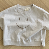 Ein weißes, kurzes T-Shirt namens Smile Tee aus der Bebe Holic-Kollektion mit einem großen Smiley auf der Vorderseite liegt auf einem hellen Holzboden mit Fischgrätenmuster. Das Smile Tee besteht aus weicher Baumwolle, hat kurze Ärmel und ein lässiges Design.