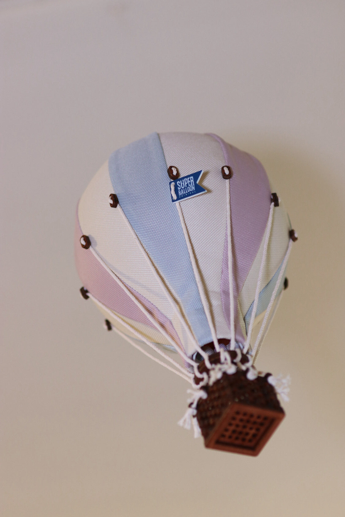 Ein kleiner dekorativer Ballon – Beige, Mint, Grau und Lila | Einzigartiges Heißluftballon-Ornament für Ihr Zuhause mit pastellblauen, weißen und rosa Paneelen. Der Korb besteht aus braunem Weidengeflecht und ist mit weißen Schnüren mit dem Ballon verbunden. Auf dem Ballon steht „SUPER 500 BALLOON“. Diese vielseitige Dekoration von Superballoon fällt vor einer schlichten, hellen Wand auf.