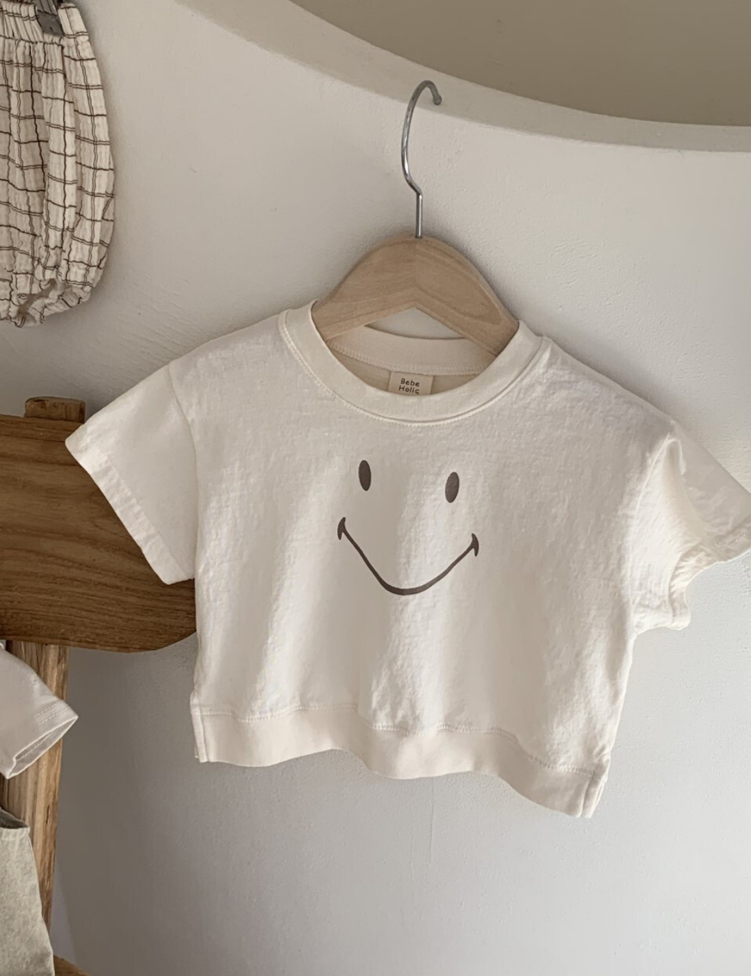 Ein minimalistisches weißes kurzes T-Shirt mit dem Namen Bebe Holic Smile Tee und einem einfachen Smiley-Design hängt auf einem hölzernen Kleiderbügel vor einer weißen Wand. Das Shirt aus weicher Baumwolle ist kurzärmlig. Daneben liegen karierter Stoff und eine Holzstufe, die eine ruhige und organisierte Atmosphäre schaffen.