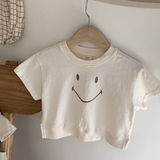 Ein minimalistisches weißes kurzes T-Shirt mit dem Namen Bebe Holic Smile Tee und einem einfachen Smiley-Design hängt auf einem hölzernen Kleiderbügel vor einer weißen Wand. Das Shirt aus weicher Baumwolle ist kurzärmlig. Daneben liegen karierter Stoff und eine Holzstufe, die eine ruhige und organisierte Atmosphäre schaffen.