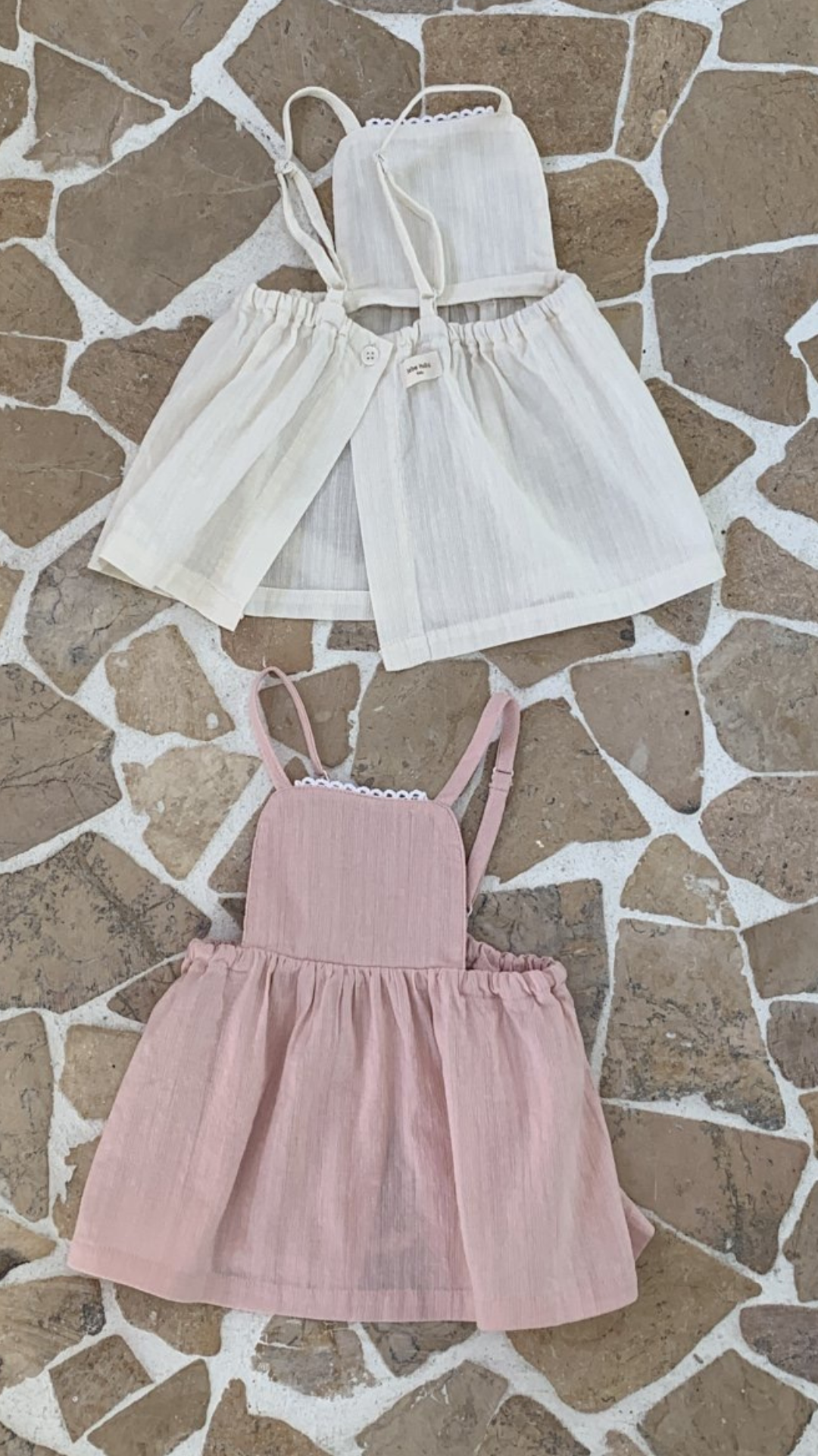 Auf einem gemusterten Steinboden sind drei Kinderkleider von Bebe Holic ausgestellt: zwei in Hellbeige und eines in Zartrosa, alle in schlichtem, elegantem Design, aus leichten Stoffen.