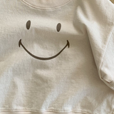 Ein kleines, hellrosa Bebe Holic Smile Tee liegt auf einem Holzboden. Das Sweatshirt hat auf der Vorderseite ein großes, einfaches schwarzes Smiley-Design und ist aus weicher Baumwolle.