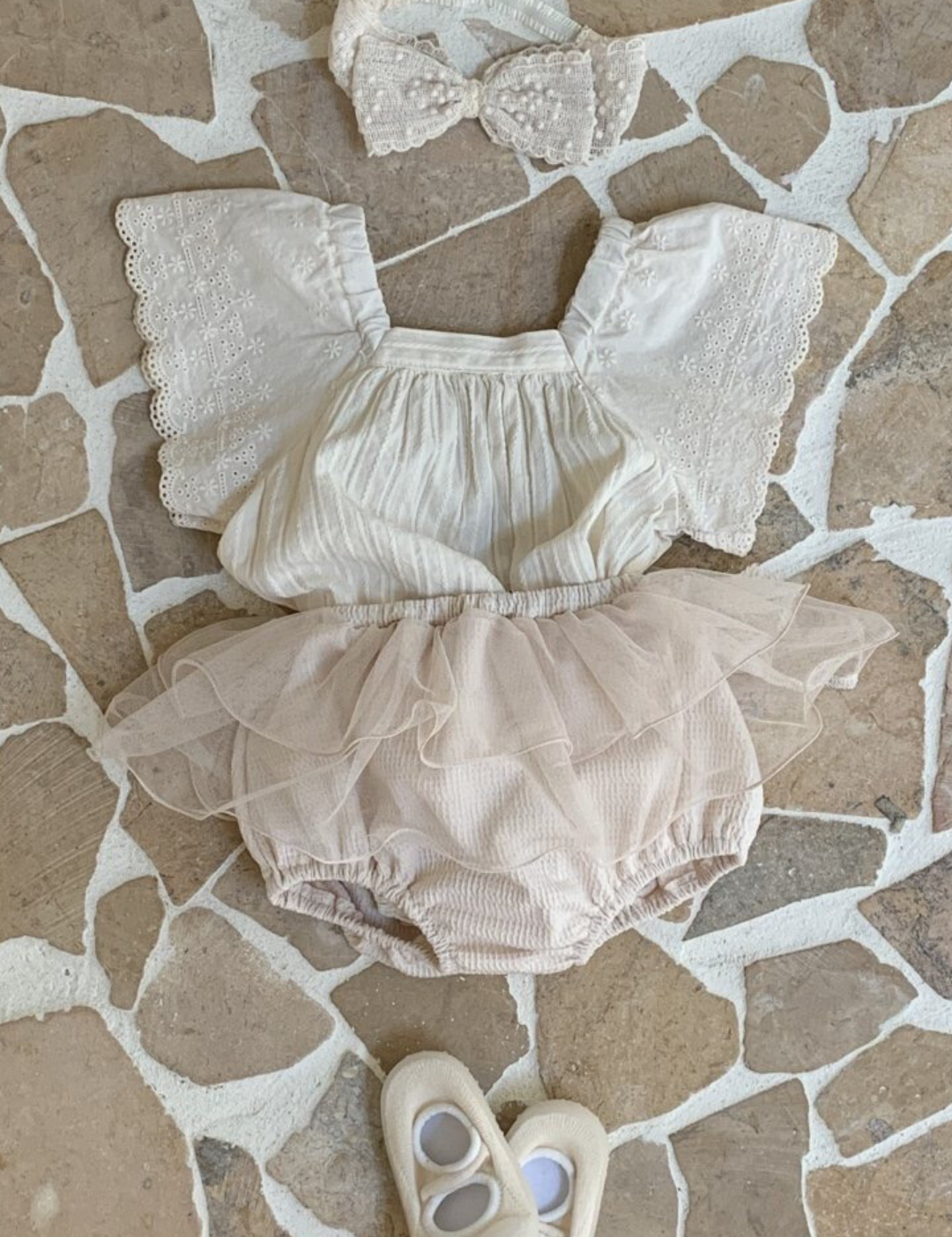 Ein Baby-Outfit, bestehend aus einem weißen Oberteil mit Spitzenärmeln, rosa Pumphosen mit Tüllüberzug und passenden weißen Schuhen, ausgebreitet auf einem gemusterten Fliesenboden.