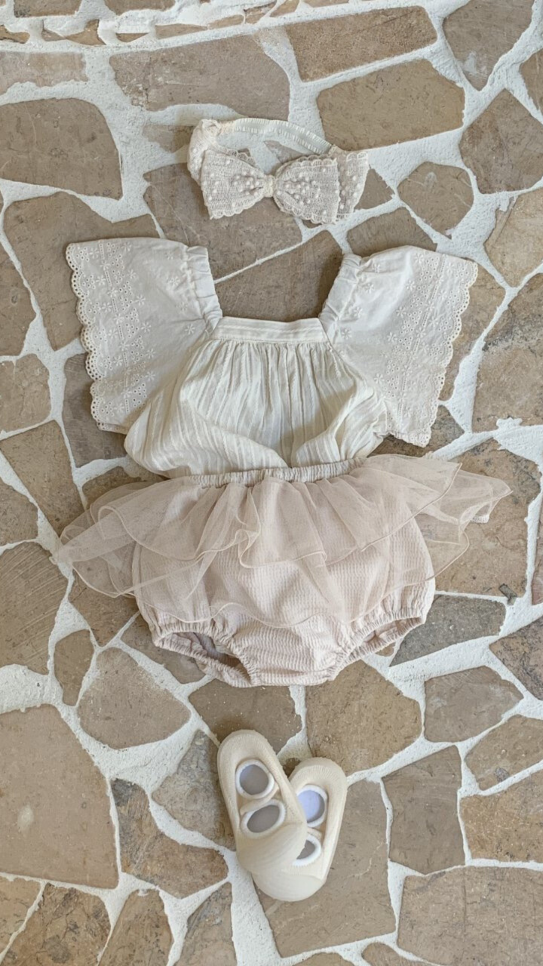 Ein Baby-Outfit, bestehend aus einem weißen Oberteil mit Spitzenärmeln, rosa Pumphosen mit Tüllüberzug und passenden weißen Schuhen, ausgebreitet auf einem gemusterten Fliesenboden.