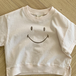 Ein blassrosa T-Shirt aus weicher Baumwolle liegt auf einem Holzboden. Das Bebe Holic Smile Tee hat in der Mitte ein einfaches schwarzes Smiley-Design. Der Boden besteht aus hellem Holz mit einem Fischgrätenmuster.