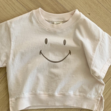 Ein blassrosa T-Shirt aus weicher Baumwolle liegt auf einem Holzboden. Das Bebe Holic Smile Tee hat in der Mitte ein einfaches schwarzes Smiley-Design. Der Boden besteht aus hellem Holz mit einem Fischgrätenmuster.