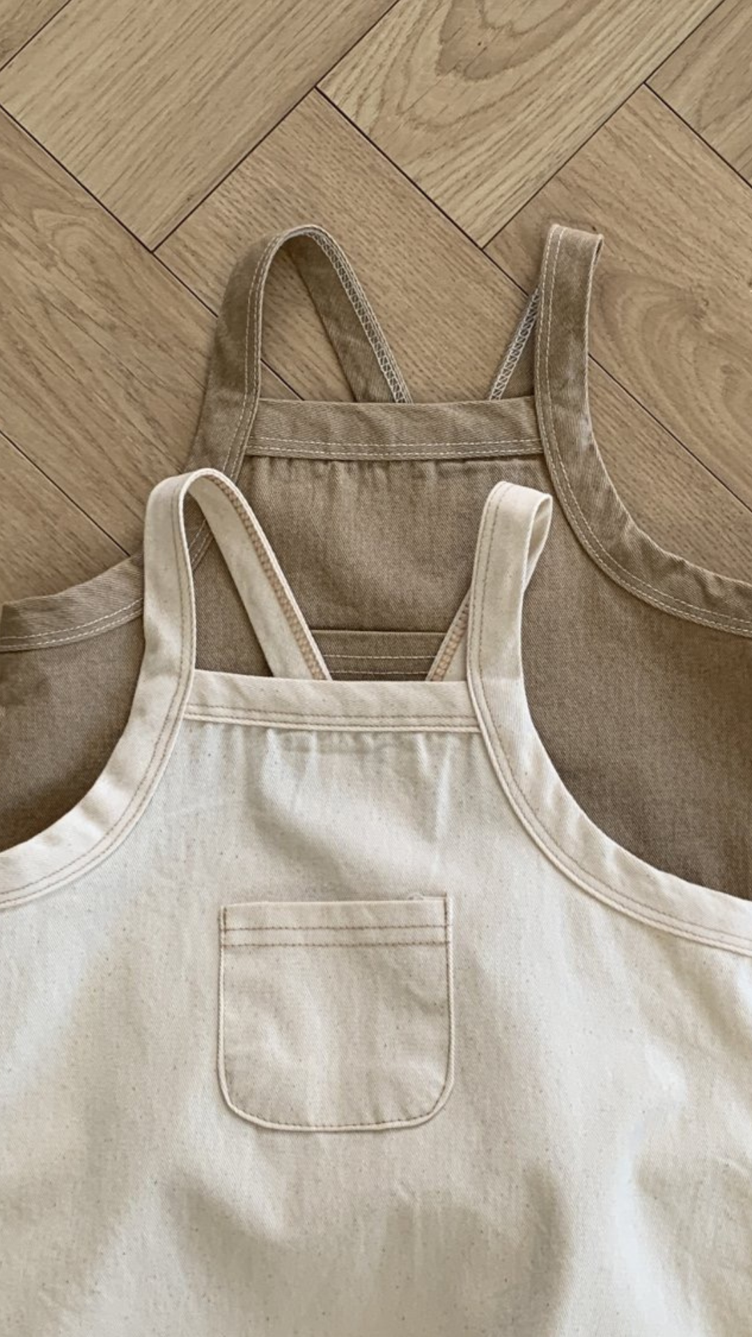 Zwei Twill-Bodysuits mit Taschen von Bebe Holic liegen auf einem hellen Fischgrätmusterboden aus Holz. Ein Bodysuit ist hellbeige mit einer kleinen Vordertasche, der andere ist dunkler beige und hat eine schlichte Vorderseite. Beide Bodysuits sind aus weicher Baumwolle gefertigt und haben verstellbare Träger für eine einfache Anpassung.