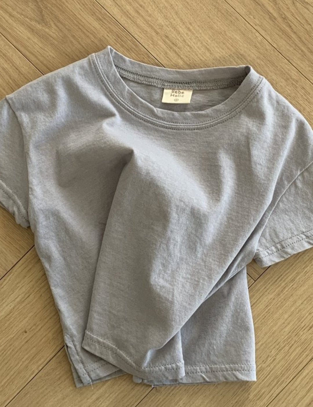 Ein hellgraues Basic-T-Shirt von Bebe Holic aus hochwertiger Baumwolle ist ordentlich gefaltet und auf einem Holzboden mit Fischgrätenmuster platziert. Das T-Shirt hat ein einfaches Design mit kurzen Ärmeln und ergänzt natürliche Töne.