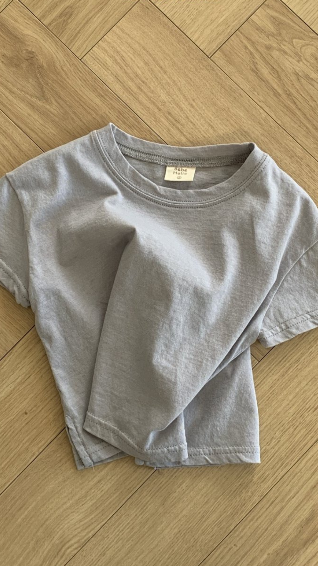 Ein hellgraues Basic-T-Shirt von Bebe Holic aus hochwertiger Baumwolle ist ordentlich gefaltet und auf einem Holzboden mit Fischgrätenmuster platziert. Das T-Shirt hat ein einfaches Design mit kurzen Ärmeln und ergänzt natürliche Töne.