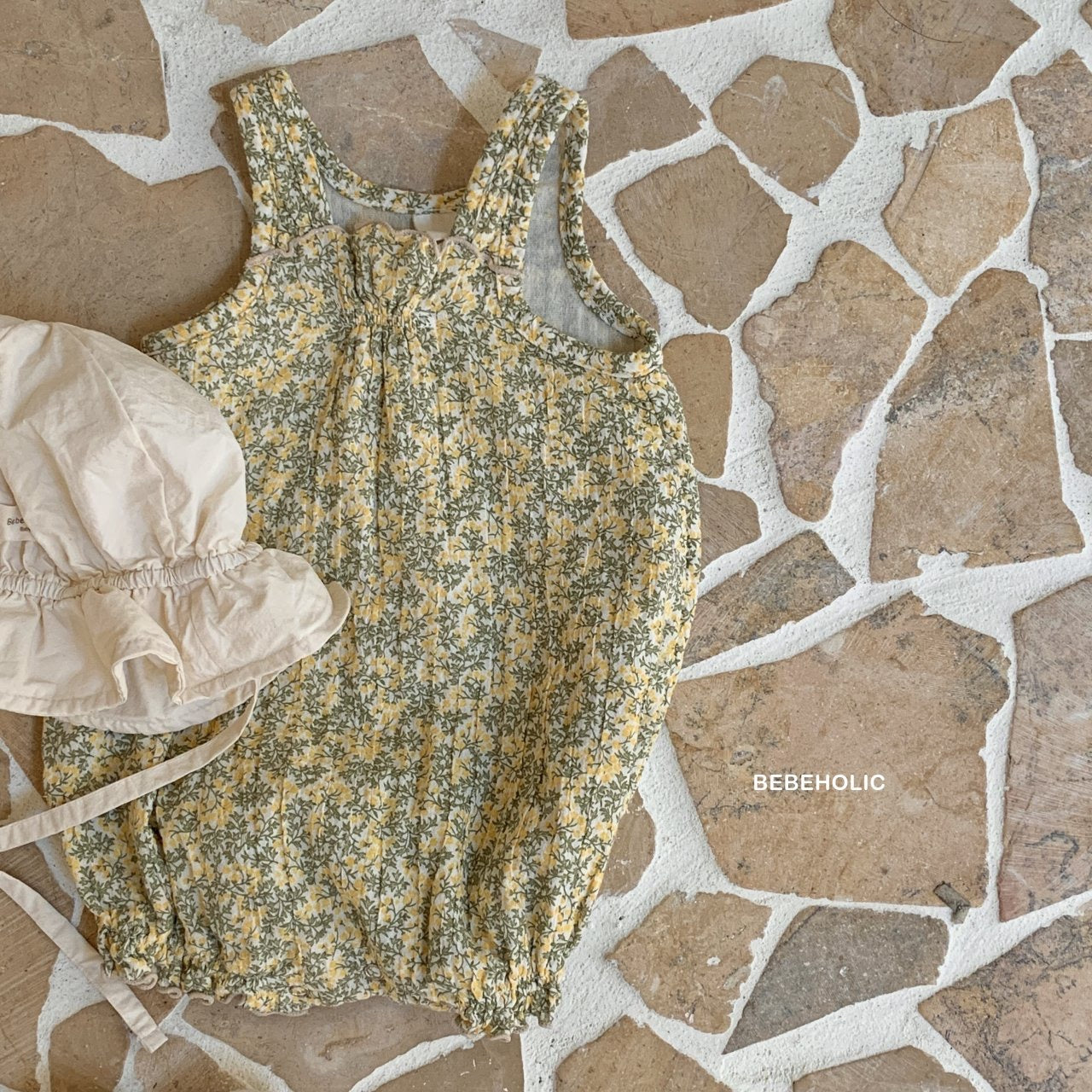 Ein gelber Karen-Bodysuit für Babys mit Blumenmuster aus atmungsaktiver Baumwolle liegt auf einer rustikalen, strukturierten Oberfläche. Daneben liegt ein cremefarbener Sonnenhut mit Kinnriemen. In der unteren rechten Ecke steht „Bebe Holic“.