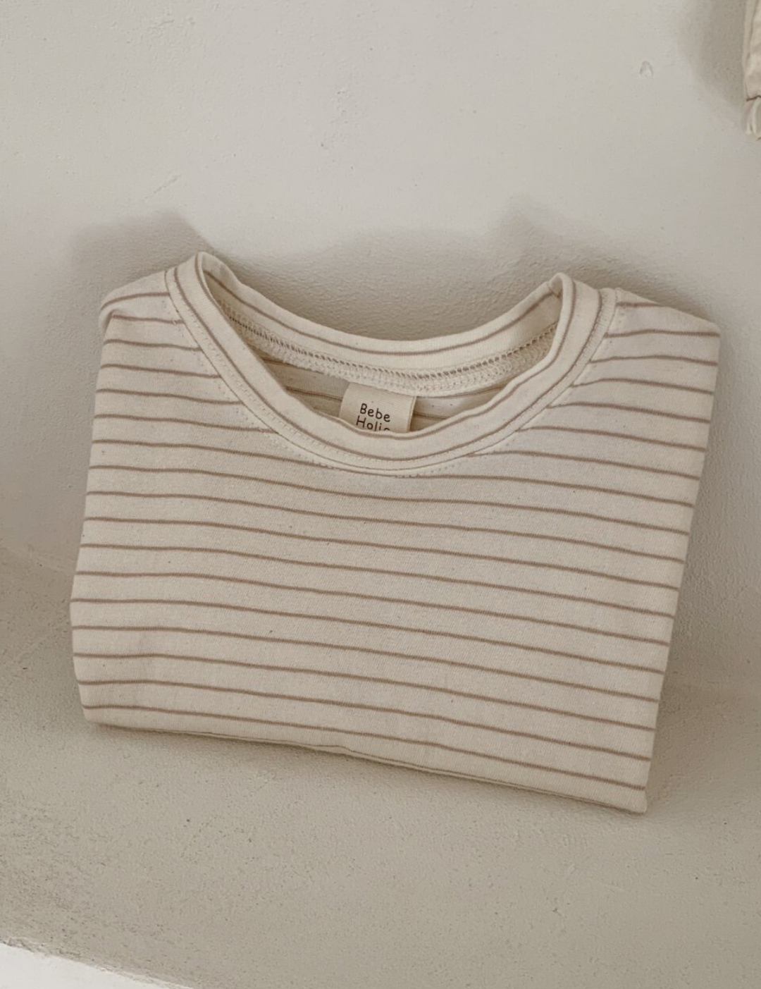 Ein ordentlich gefaltetes, beige-weiß gestreiftes Langarmshirt mit der Aufschrift „Basic ST Tee“ von Bebe Holic liegt auf einer weißen Fläche. Dieses gestreifte Baby-T-Shirt verströmt klassischen Stil, wobei sein schlichter, heller Hintergrund die Einfachheit und Sauberkeit des Designs unterstreicht.