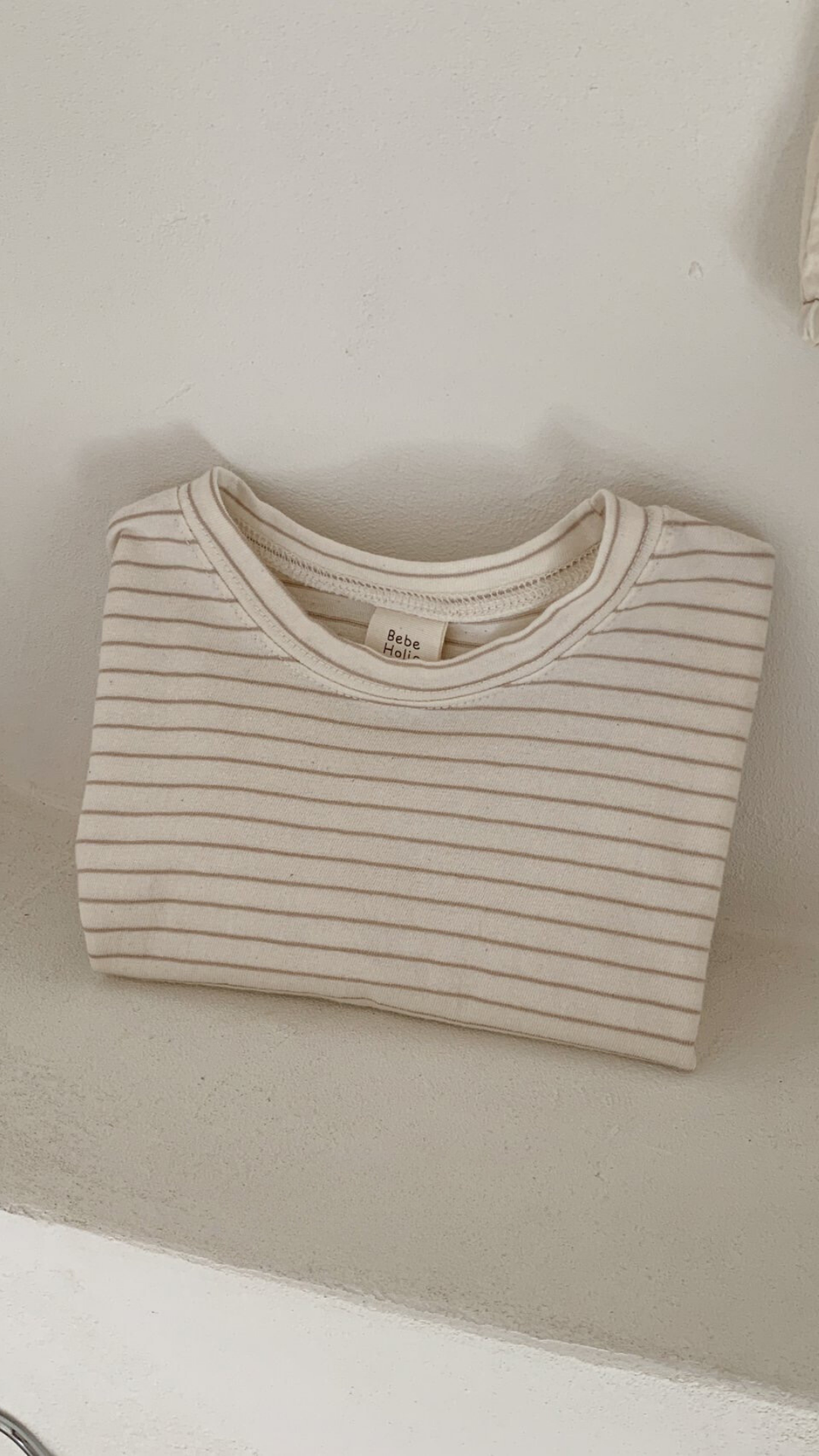 Ein ordentlich gefaltetes, beige-weiß gestreiftes Langarmshirt mit der Aufschrift „Basic ST Tee“ von Bebe Holic liegt auf einer weißen Fläche. Dieses gestreifte Baby-T-Shirt verströmt klassischen Stil, wobei sein schlichter, heller Hintergrund die Einfachheit und Sauberkeit des Designs unterstreicht.