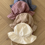 Fünf Crunch Bucket Hats in einer natürlichen Farbpalette (Blau, Rosa, Braun, Beige und Creme) sind ordentlich auf einem hölzernen Boden im Fischgrätenmuster gestapelt. Der cremefarbene Hut oben hat zwei dünne Riemen. Entdecken Sie die bezaubernde Auswahl von Bebe Holic für Ihre Kleinen.