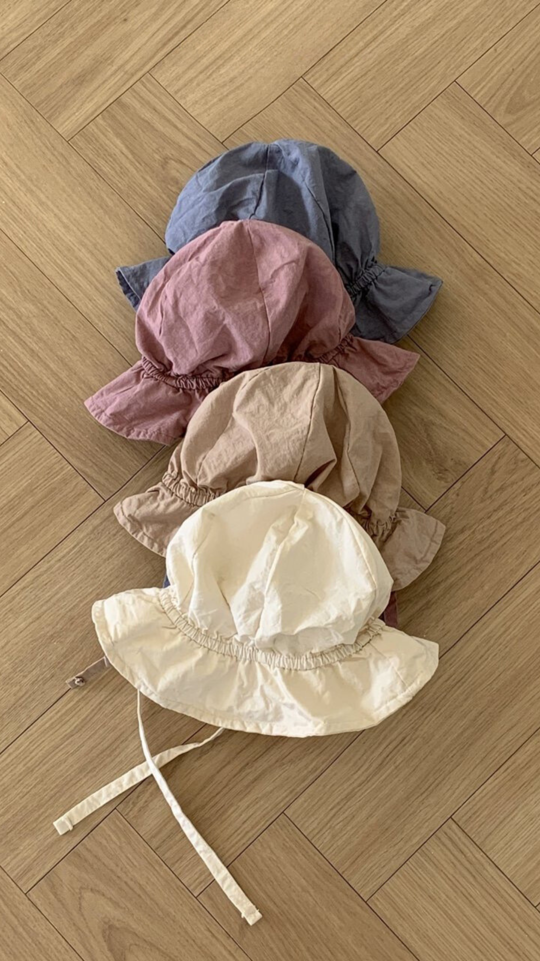 Vier Crunch Bucket Hats in verschiedenen gedeckten Tönen (Blau, Mauve, Beige und Creme) von Bebe Holic sind ordentlich auf einem hellen Holzboden mit Fischgrätenmuster gestapelt, wodurch ihre natürliche Farbpalette hervorgehoben wird.