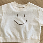 Ein weißes, kurzes Sweatshirt aus weicher Baumwolle mit einer einfachen, großen Smiley-Grafik auf der Vorderseite. Das Smile Tee von Bebe Holic liegt flach auf einem hellen Holzboden im Fischgrätenmuster.