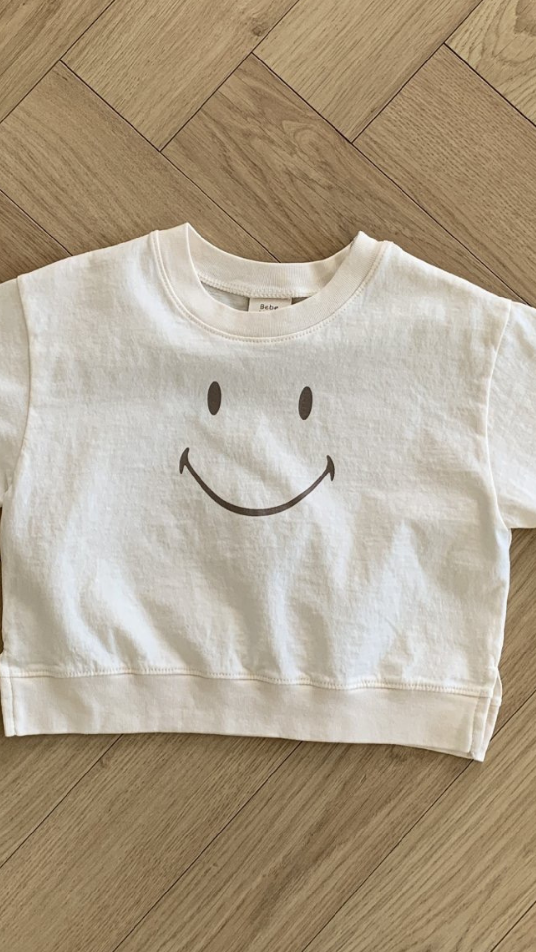 Ein weißes, kurzes Sweatshirt aus weicher Baumwolle mit einer einfachen, großen Smiley-Grafik auf der Vorderseite. Das Smile Tee von Bebe Holic liegt flach auf einem hellen Holzboden im Fischgrätenmuster.