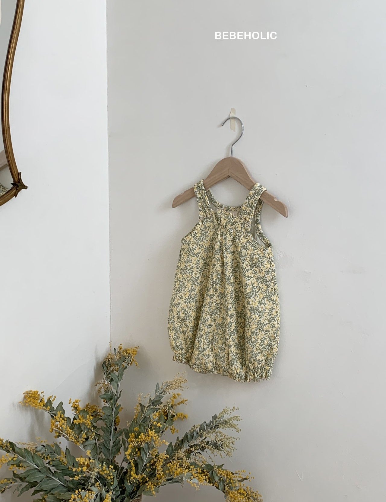 Ein Karen Body von Bebe Holic aus atmungsaktiver Baumwolle hängt auf einem hölzernen Kleiderbügel vor einer hellgrauen Wand. Das verspielte Outfit ist mit kleinen Blumen in Gelb- und Grüntönen geschmückt. Ein kleiner Strauß gelber Blumen liegt darunter und verleiht der Szene einen Hauch von natürlichem Charme.