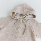 Ein beigefarbener Baby-Hoodie mit Druckknöpfen und Kapuze liegt flach auf einer weißen Oberfläche. Der Hoody-Bodysuit hat Kordeln an der Kapuze und ein Markenlabel „Bebe Holic“ ist unter den Knöpfen auf den Stoff genäht, um Babykomfort für Ihren Kleinen zu gewährleisten.