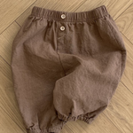 Ein Paar braune Shorts in Kleinkindergröße mit elastischer Taille und zwei Knöpfen auf der Vorderseite. Die leichten Bebe Holic Natural Pants haben elastische Bündchen an den Beinöffnungen. Diese vielseitigen Must-have-Shorts werden flach auf einem Holzboden ausgebreitet.