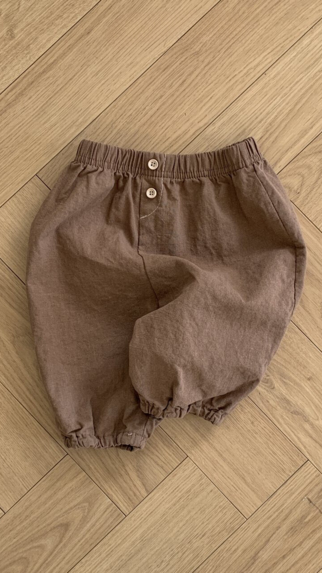 Ein Paar braune Shorts in Kleinkindergröße mit elastischer Taille und zwei Knöpfen auf der Vorderseite. Die leichten Bebe Holic Natural Pants haben elastische Bündchen an den Beinöffnungen. Diese vielseitigen Must-have-Shorts werden flach auf einem Holzboden ausgebreitet.