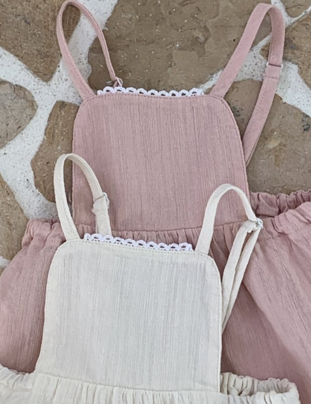 Drei pastellfarbene Bebe Holic-Kinderkleider mit Rüschenträgern und Spitzendetails, vertikal auf einem strukturierten Steinhintergrund angeordnet. Diese aus leichten Stoffen gefertigten Sommerkleider sind perfekt für warmes Wetter.