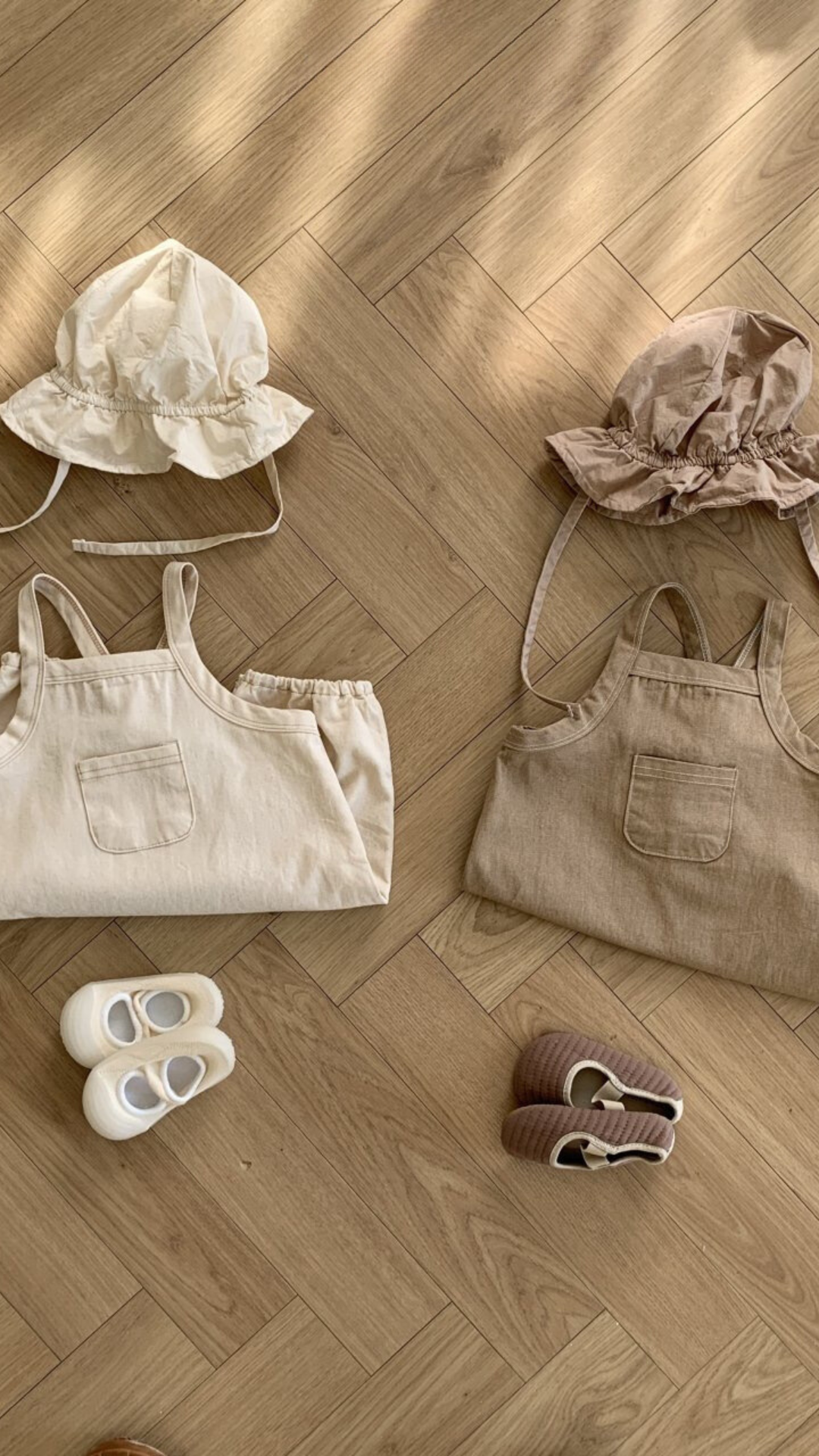 Zwei Sets mit Kleinkinder-Outfits liegen auf einem Holzboden. Jedes Set enthält einen Twill-Body mit verstellbaren Trägern, eine Haube und ein Paar Schuhe. Das linke Set ist cremefarben mit passenden Schuhen und aus weicher Baumwolle gefertigt, und das rechte Set ist beige mit braunen Schuhen. Sonnenlicht fällt durch und wirft weiche Schatten. Diese entzückenden Ensembles sind von Bebe Holic.