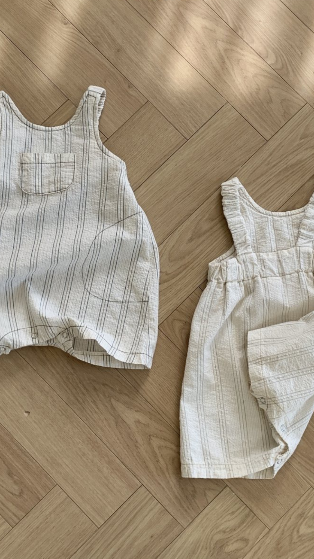 Zwei Baby-Outfits auf einem Holzboden; eines ist ein graues ärmelloses Oberteil mit passenden Shorts, das andere ist ein cremefarbener Bebe Holic Ariel St Body mit geraffter Taille.