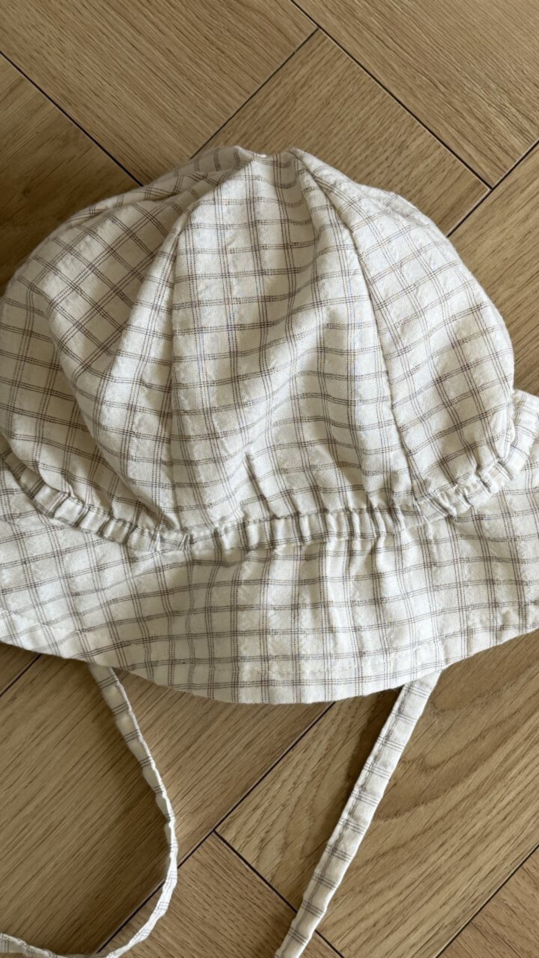 Ein beige karierter Bucket Hat mit breiter Krempe und Kinnriemen, der an traditionelle Karo-Kinderhüte erinnert, liegt flach auf einem Holzboden. Der Stoff hat ein weiches, gitterartiges Muster mit hellbraunen Linien für optimalen Sonnenschutz. Die Krempenränder sind leicht gebogen. Dies ist der Yogurt ST Bucket Hat von Bebe Holic.