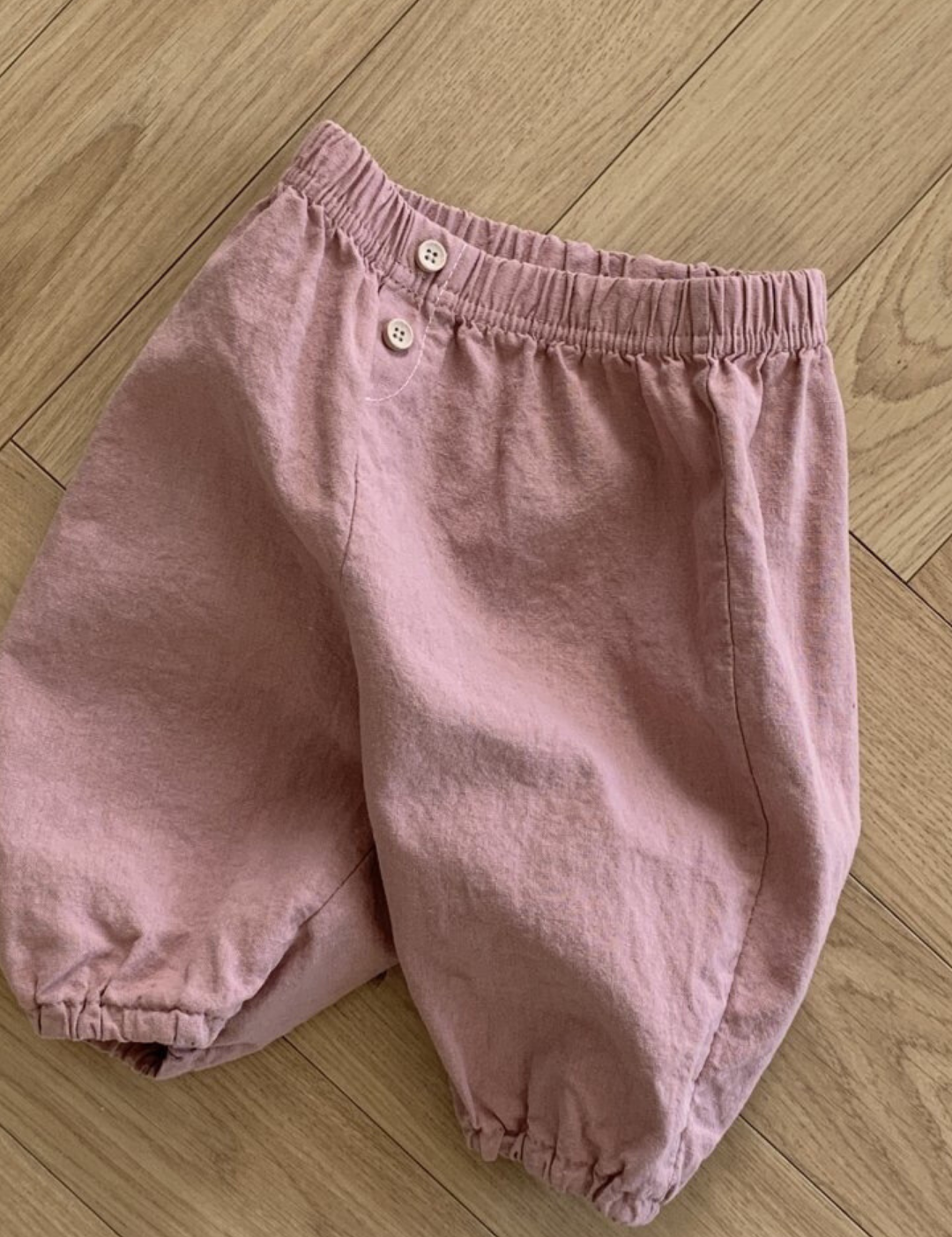 Ein Paar hellrosa Bebe Holic Natural Pants aus zerknittertem Stoff mit elastischem Bund und zwei Knöpfen vorne. Diese vielseitigen Must-have-Shorts haben lockere, geraffte Säume und werden flach auf einen Holzboden gelegt.