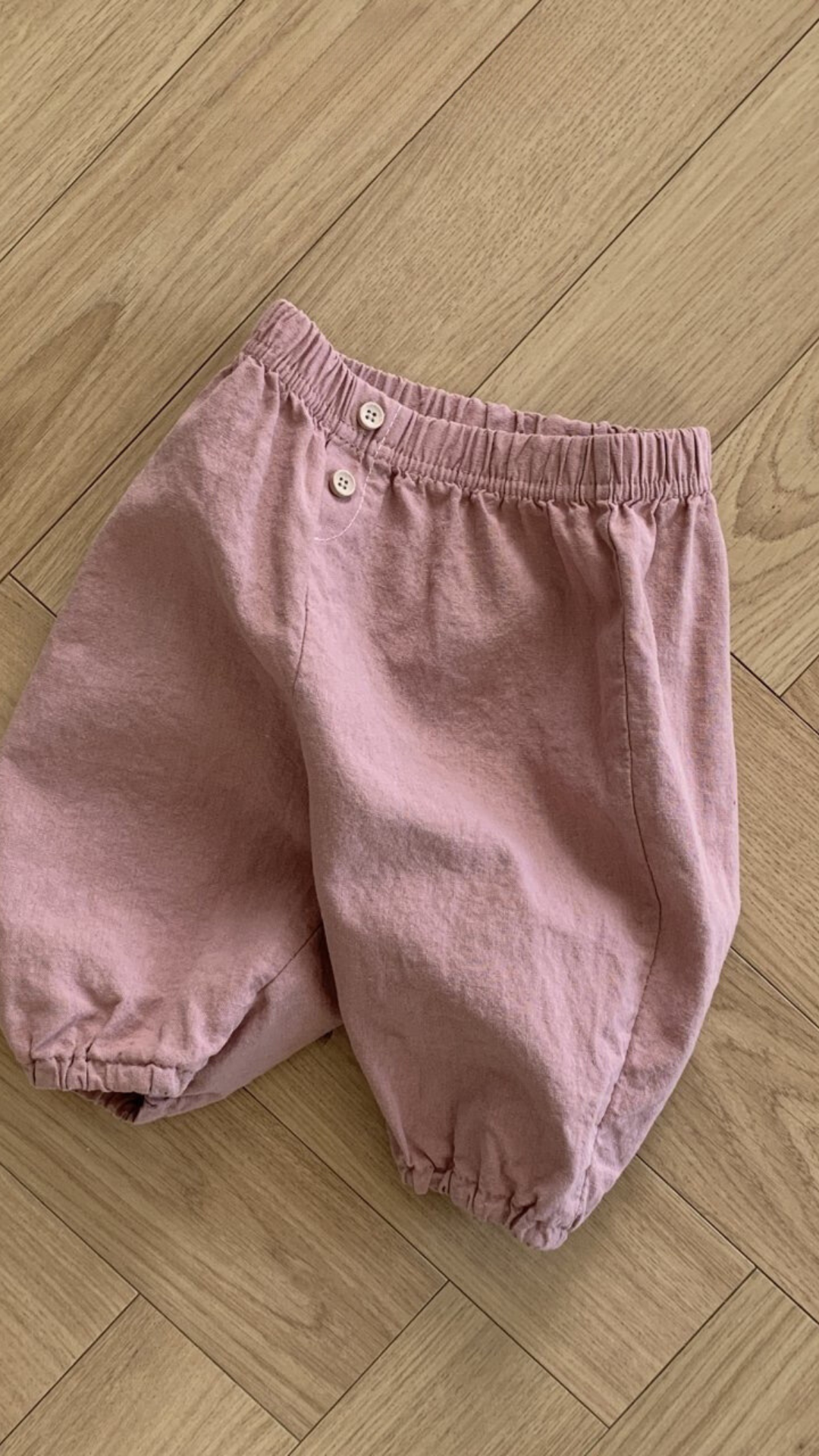 Ein Paar hellrosa Bebe Holic Natural Pants aus zerknittertem Stoff mit elastischem Bund und zwei Knöpfen vorne. Diese vielseitigen Must-have-Shorts haben lockere, geraffte Säume und werden flach auf einen Holzboden gelegt.