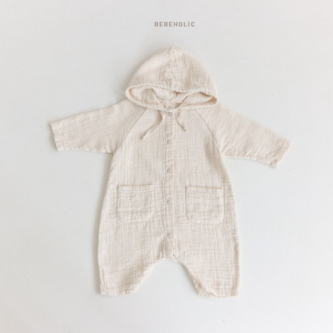 Ein beiger Baby-Strampler mit langen Ärmeln, Kapuze und Vordertaschen auf weißem Hintergrund. Der Strampler hat einen strukturierten Stoff und eine Kordel an der Kapuze. Der Text oben lautet „Bebe Holic Hoody Bodysuit“.